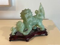 Side view of green jade Foo dog figurine on a wooden base, showing the smaller Foo dog at the back and the overall shape of the larger dog.