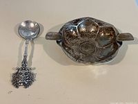 Photo showing both Brazilian sterling silver spoon and ashtray together on a white surface, spoon has coin bowl and detailed handle, ashtray shows scalloped design and small handles.