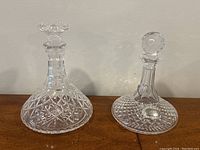 Two clear crystal decanters side by side on wooden surface; left one flat topped marked Stuart Crystal, right one with rounded stopper; silver plated Port label in front