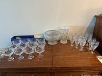 Full view of cut crystal rose bowl, flower frog, large vase, multiple sizes of stemmed drinking glasses, and large candle stick holder on wooden table.