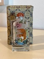 Front view of crystal scent bottle showing detailed hand painted tiger and nature scene inside, stone stopper on top, and original patterned box in background.