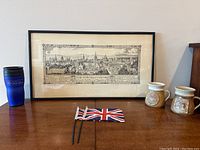 Framed antique print of The Towers of Oxford, blue travel mug, two University of Newcastle pottery mugs, and pair of small Union Jack flags displayed on wooden surface against wall.