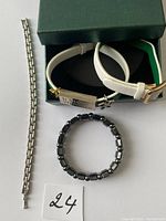 Lot of 3 bracelets arranged with the white watch placed in a green Shaklee box, silver-tone metal link bracelet laid flat beside the box, and black hematite bead stretch bracelet coiled beneath the watch box. The watch has a rectangular silver-tone case and a price tag attached.