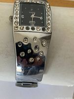 Close-up of silver-tone fashion bracelet watch with embedded crystals and black face showing brand and quartz movement.