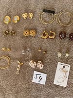 Multiple pairs of various style earrings including gold-tone hoops, studs, pearl clusters, and colored stones all arranged on a gray textured surface with lot number card.