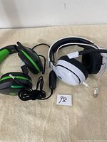 Photo showing two wired over-ear headphones; one black and green with mic and USB connector, the other white and black with padded ear cups and cable control.