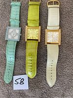 Full front view of three Joan Rivers Classics watches laid flat on carpet with paper marked 58. Colors are pastel light green, lime green, and off-white with square watch faces and gold-tone bezels.