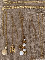 Lot of 12 necklaces displayed on a beige fabric surface, showing variety in chain styles and pendants including beaded, metal, and leaf motifs