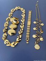 Three gold-toned vintage jewelry pieces photographed on a blue background: two necklaces (one chunky link chain with hearts and ovals, one chain with small discs) and a bracelet watch with rectangular face.