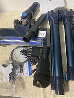 Photo showing the main vacuum cleaner unit, two extension tubes, motorized floor brush, various accessories including crevice tool, round brush, charger, and additional plastic attachment on a light surface.