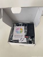 Open box showing LED strip light components, power adapter, and instruction manual with colorful circular LED illustration.