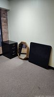 Photo of a black metal 3-drawer file cabinet, an octagonal wooden framed mirror, and a black square table top against a wall on carpeted floor.