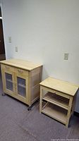 Two wooden storage cabinets against wall, one larger with glass doors and wheels, one smaller with open shelves