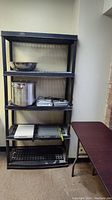 Black plastic shelving unit with five shelves holding stainless steel bowl, large pot, electric double hot plate, trays.