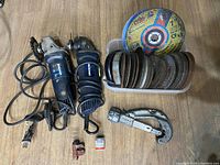 Wide view showing two electric angle grinders, one with a wire wheel brush attachment and one with a cutting disc attached. Surrounding them is a plastic bin with multiple cutting and grinding discs, a small red pipe cutter tool, and a Dremel accessory container.