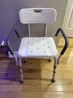 Front view of white shower chair with backrest, black padded armrests, drainage holes in seat, and adjustable metal legs ending in rubber tips, placed on wooden floor in front of wall.