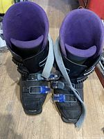 Front top view of pair of black Tecnica ski boots with purple interior lining and front buckles on wooden floor.