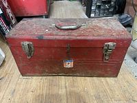 Closed red metal toolbox showing worn paint, dents, and a handle on top with two latches securing the lid.