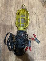 Full view of yellow work light with metal cage and attached black cord with clamps, placed on wooden surface.