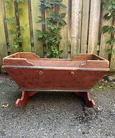 Side view of the antique wooden cradle showing painted red trapezoidal body and curved rocker base on outdoor surface against fence and plants.