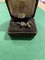 Four gold tone vintage rings displayed on black and green surfaces with clear faceted stones and textured bands.