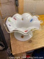 Photo showing the white opaque Murano glass bowl with colorful inclusions and clear folded wavy edges trimmed in gold, placed on a wood surface.