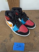 Pair of multicolor Air Jordan 1 Mid sneakers on a brown surface, showing the front angled view with visible red, blue, white, black, orange and purple leather panels.