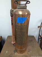 Front view of bronze fire extinguisher showing handle, hose, and embossed instruction plaque with blue lot number sticker.