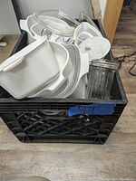 Top view of the lot showing stacked white ceramic casserole dishes with handles and clear glass lids inside a black crate.