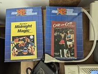 Two boxed Atari XE video game cartridges titled 'David's Midnight Magic' and 'One-On-One Basketball' with visible Atari XE logo and grid blue box pattern, along with an Atari system cable
