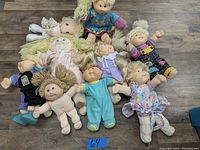 Eleven dolls shown spread out on a wood floor, mainly Cabbage Patch Kids with different outfits and hairstyles, with one Precious Moments doll in cream dress at top left.
