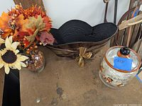 Photo showing artificial autumn floral arrangement, black decorative basket with gold bows, and glass Halloween treat jar on rustic surface.