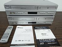 Front view of two Memorex DVD/VCR combo units stacked, showing front panel details, LCD display, buttons, and input jacks. Two remotes and user guides placed in front on surface.