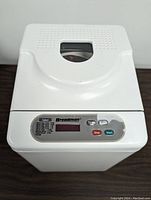 Top view of white Breadman bread machine showing closed lid with viewing window and digital control panel with buttons for select, time, stop, start.