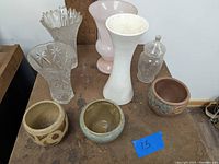 Front view of all seven items arranged on wooden surface, showing two crystal glass vases, white ceramic vase, pink glass vase, two ceramic planters, and glass jar.