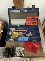 Open blue plastic box containing hammer, chisel, socket sets, drill bits, gauge, and hand saw.