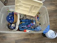 Top view of clear plastic tote with assorted party supplies inside including a blue glass pitcher, woven basket, white plastic trays, and cocktail stirrers.