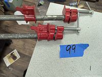Two pipe clamps on steel pipes with red cast iron jaws and screw handles laid side by side on a white surface.
