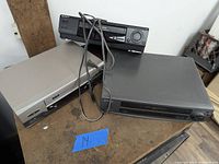 Three VCR players stacked on a wooden surface: silver RCA, dark gray Sharp, and black unit behind with cables.