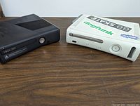 Two Xbox 360 consoles on a wooden surface, one black and one white with multiple stickers on top.