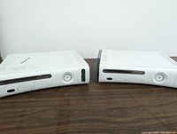 Two white Xbox 360 consoles side by side on a white surface, showing top and front sides