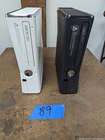 Two Xbox 360 consoles side by side on table, white and black models visible from front view with disc trays and power buttons.