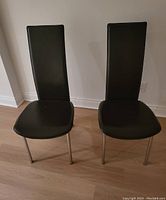 Pair of black high back chairs with leather-like upholstery and metal legs shown from front on wood floor with white wall background.