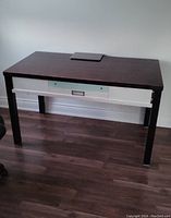 Front view of wood desk with one drawer, dark brown top, and black legs on wood floor in front of white wall.