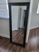 Full-length dressing mirror with a dark wood frame standing on a wooden floor, against a wall, visible window and door reflections.