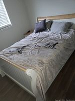 Full bed shown with mattress and bedding including pillows, fitted sheets, and comforter. Bed frame is wooden with white panels and light wood top rail on headboard and footboard.