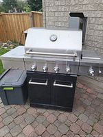 Photo showing front view of KitchenAid propane BBQ grill with stainless steel lid, built-in temperature gauge, four burners, black cabinet with double doors, and side shelves.