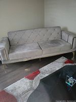 Beige upholstered two-seat sofa with button tufted backrest and armrests, metal legs, situated on wooden floor next to a wall and a rug.