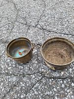 Two tarnished brass bowls placed on asphalt, one bowl has small handles and a greenish patina inside, the other is larger and without handles.