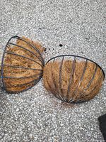 Two black metal half-moon shaped planters with brown coconut fiber liners on gravel surface.